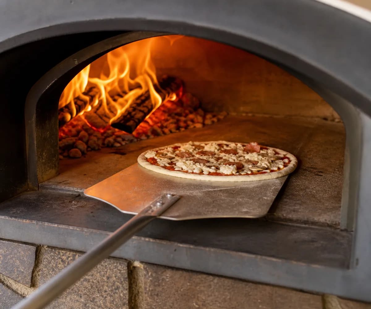 Pizza being launched from a peel onto a pizza oven floor away from the strongest flame.