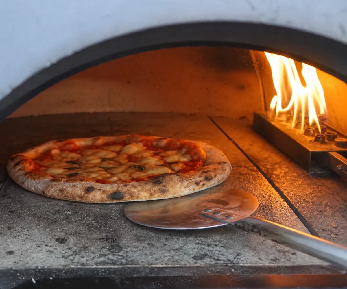 Pizza being turned with a turning peel inside a pizza oven near a steady flame.